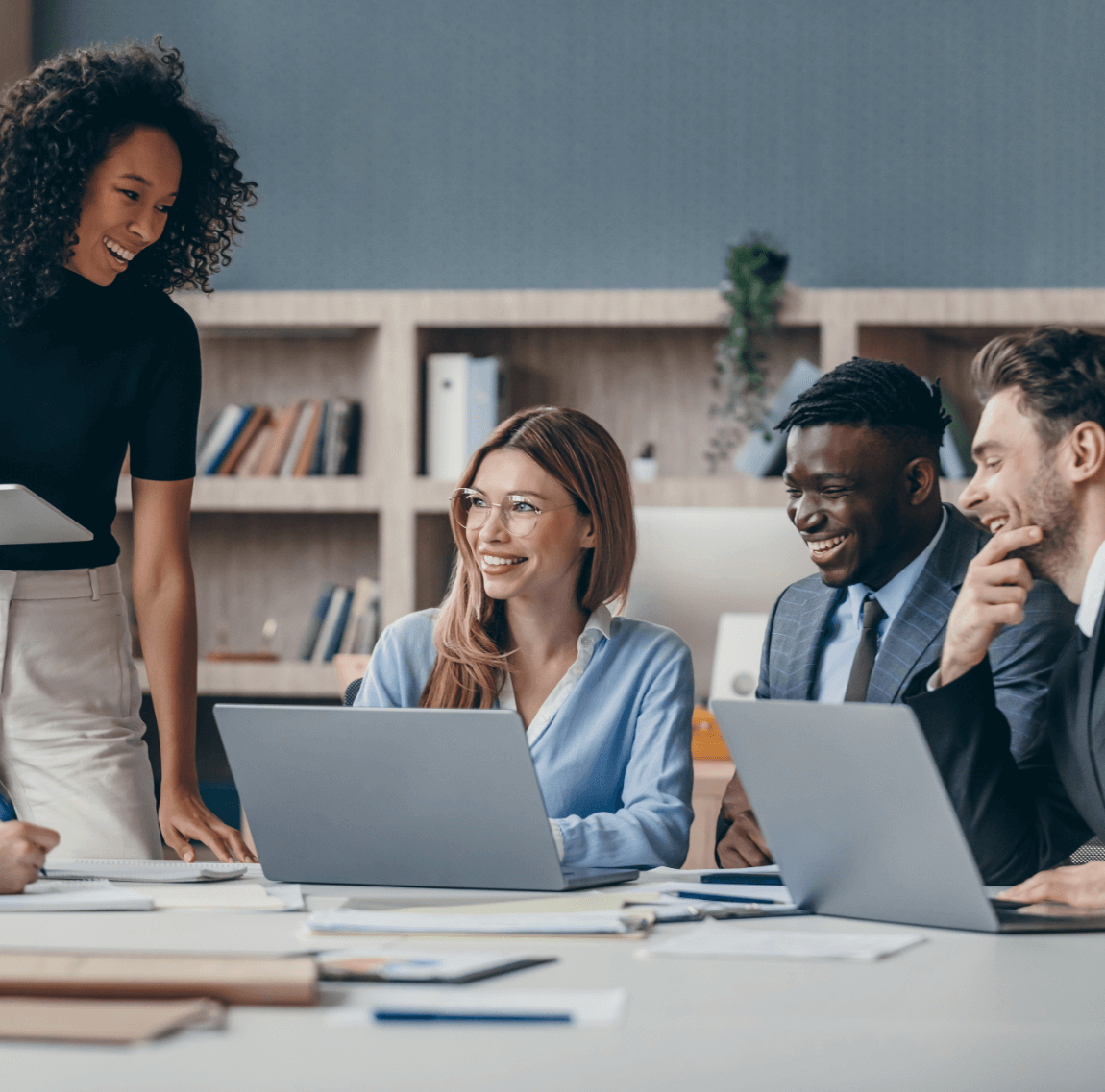 Four professionals at desk with laptops Four business professionals sitting around a desk with laptops and paperwork. A bookshelf with business books and binders in the background.