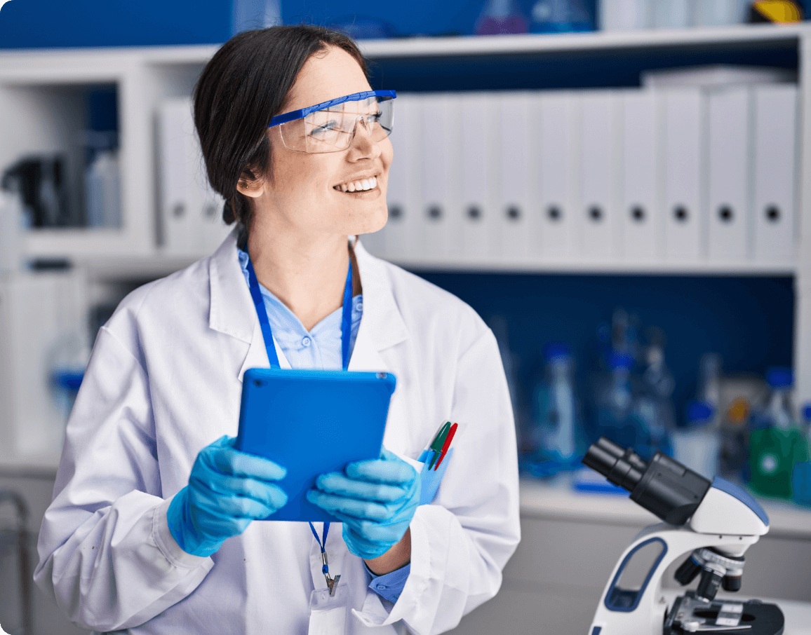 Lab technician with tablet A female lab technician smiling and holding a blue electronic tablet. A microscope on a desk nearby and shelving with binders in the background.