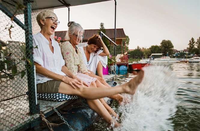 Three women sitting together at a harbor with their feet in the water. One woman is playfully kicking the water.