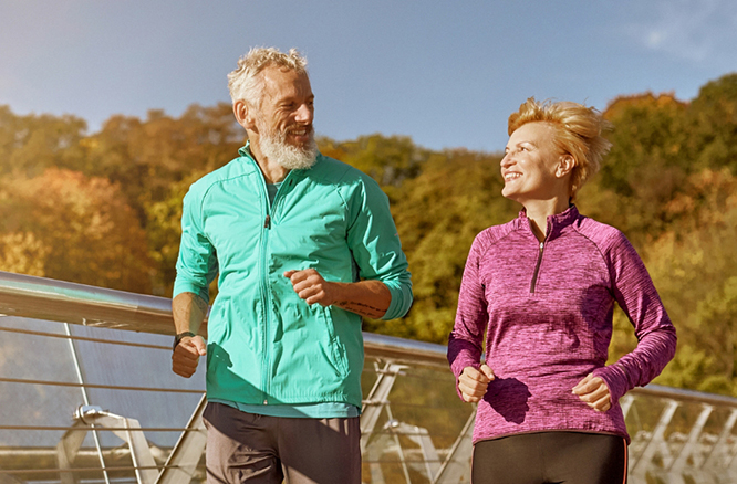 Man in teal jacket and woman in pink jacket running together across a bridge while looking toward each other.