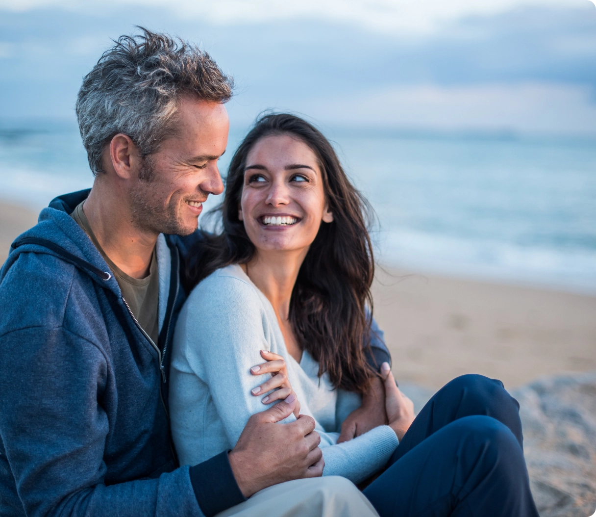 A happy couple sitting on the beach smiling and looking at each other.