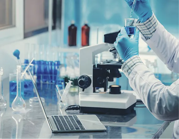 Lab technician handling a liquid sample beside a microscope and laptop, representing ingredient testing and research. 